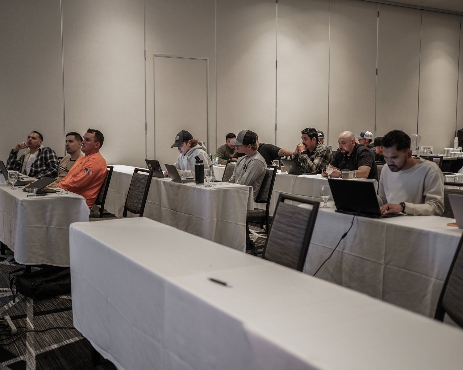 Men sitting at tables with laptops and beverages in a conference room, some engaged with their devices, while others are listening or in thought.
