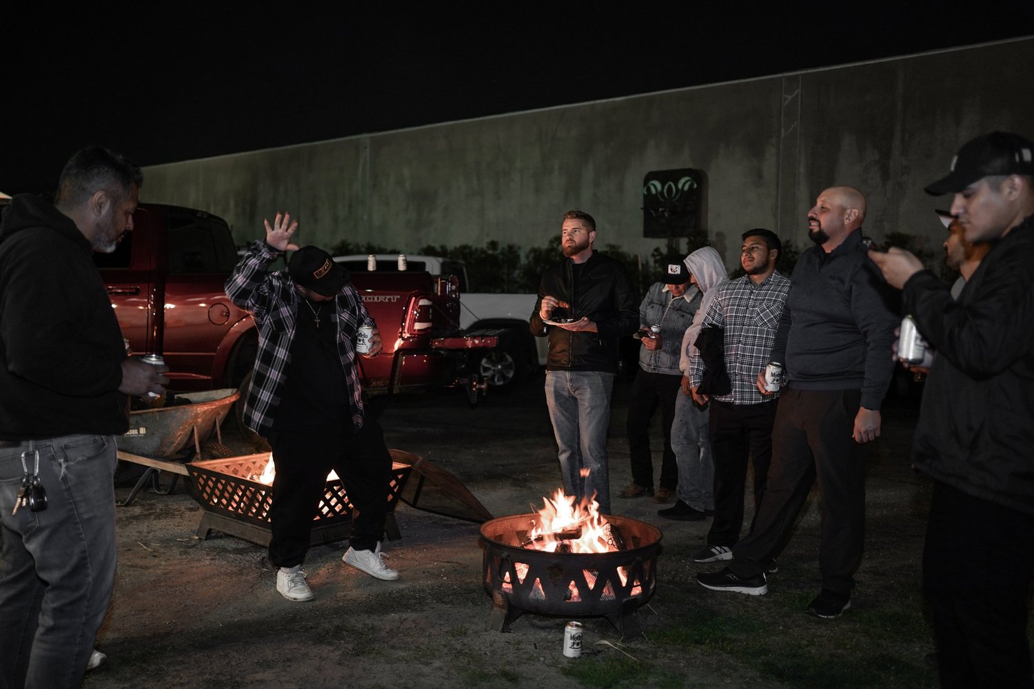 Group of people gathered around a fire at night in a parking lot, some with drinks, some looking at their phones.