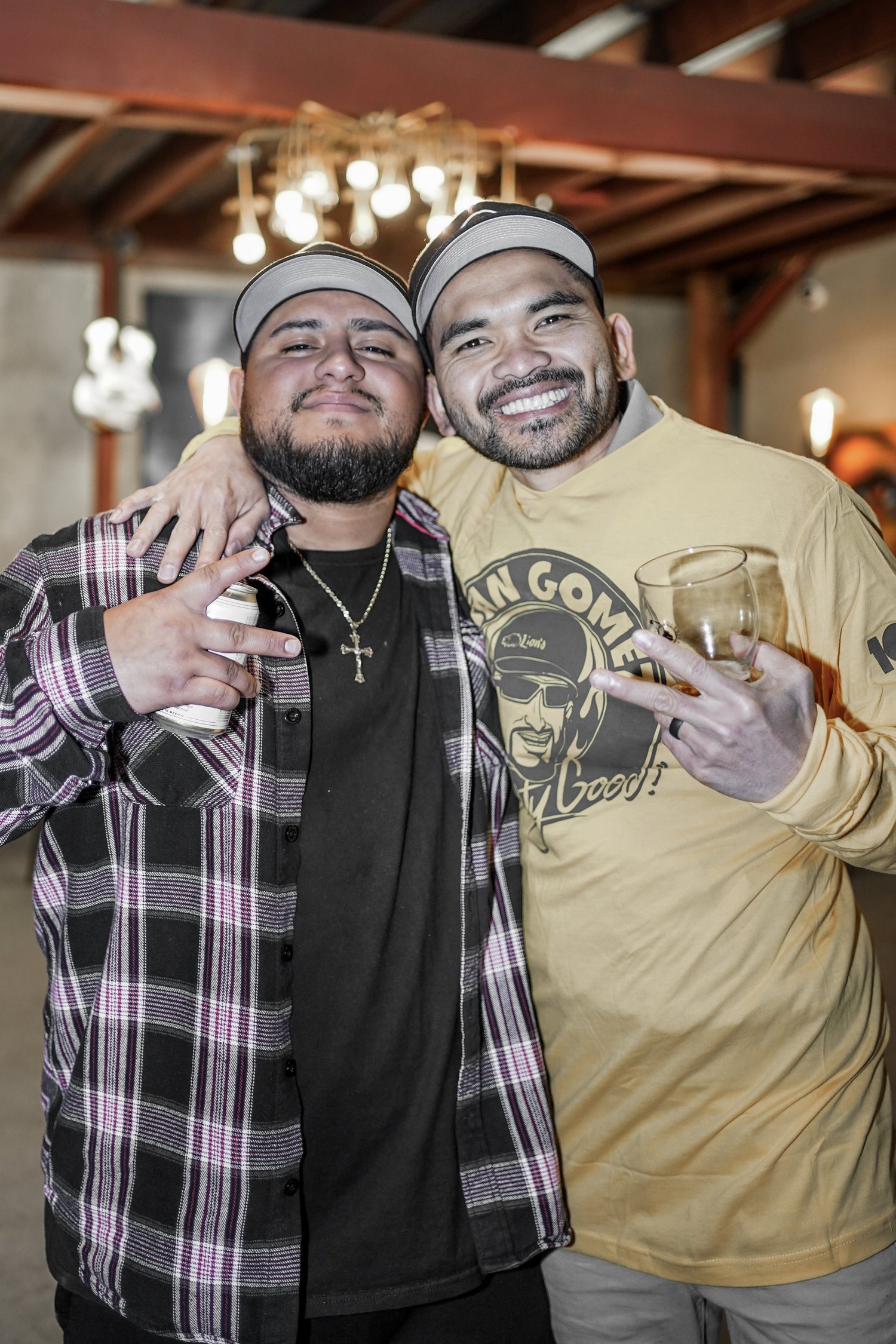 Two men smiling and posing for the camera at a social gathering, one wearing a plaid shirt and a cross necklace, the other wearing a yellow long sleeve t-shirt with a graphic print, both wearing caps.