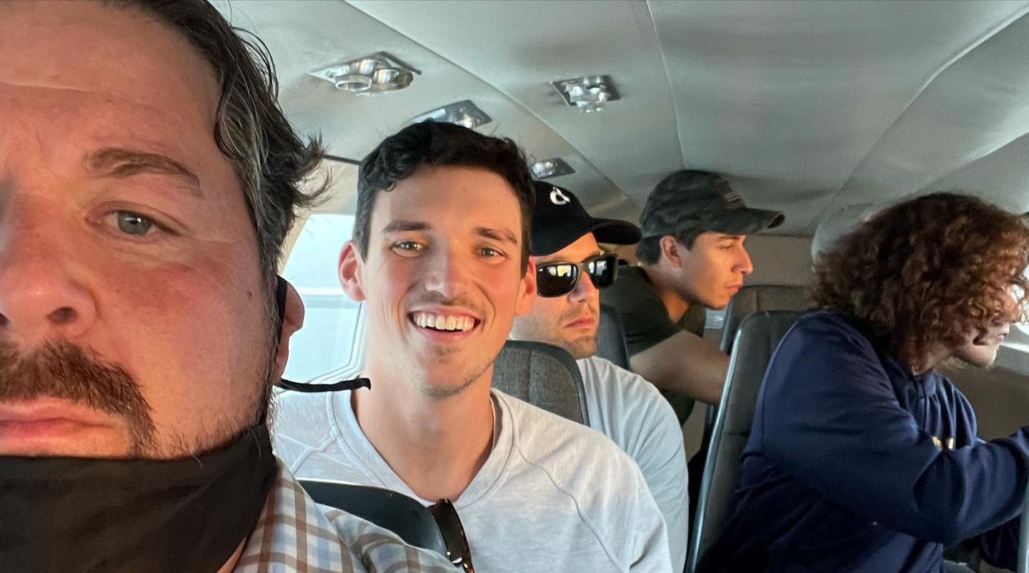 Group of five men seated inside an aircraft, some wearing sunglasses, with one man smiling at the camera and others focused on their activities.
