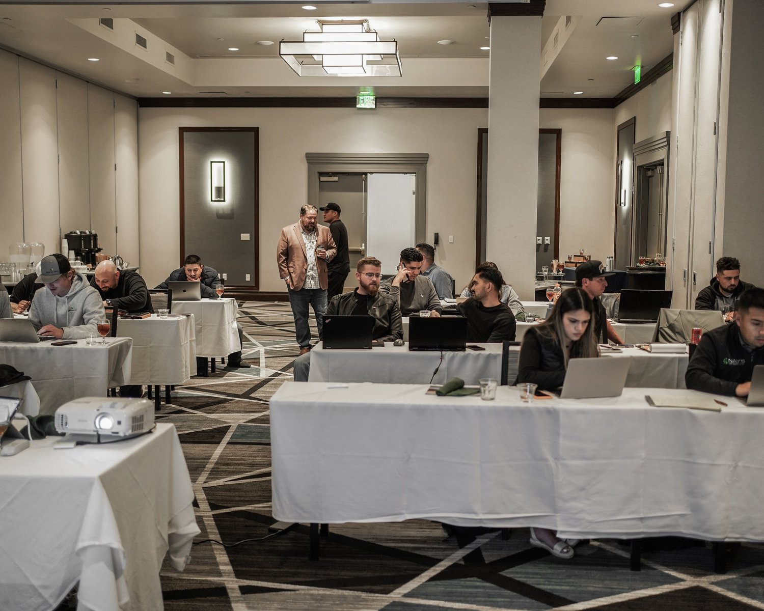 A group of people sitting at tables in a conference room, working on laptops and engaging in conversation, with two individuals standing and talking near the back of the room.