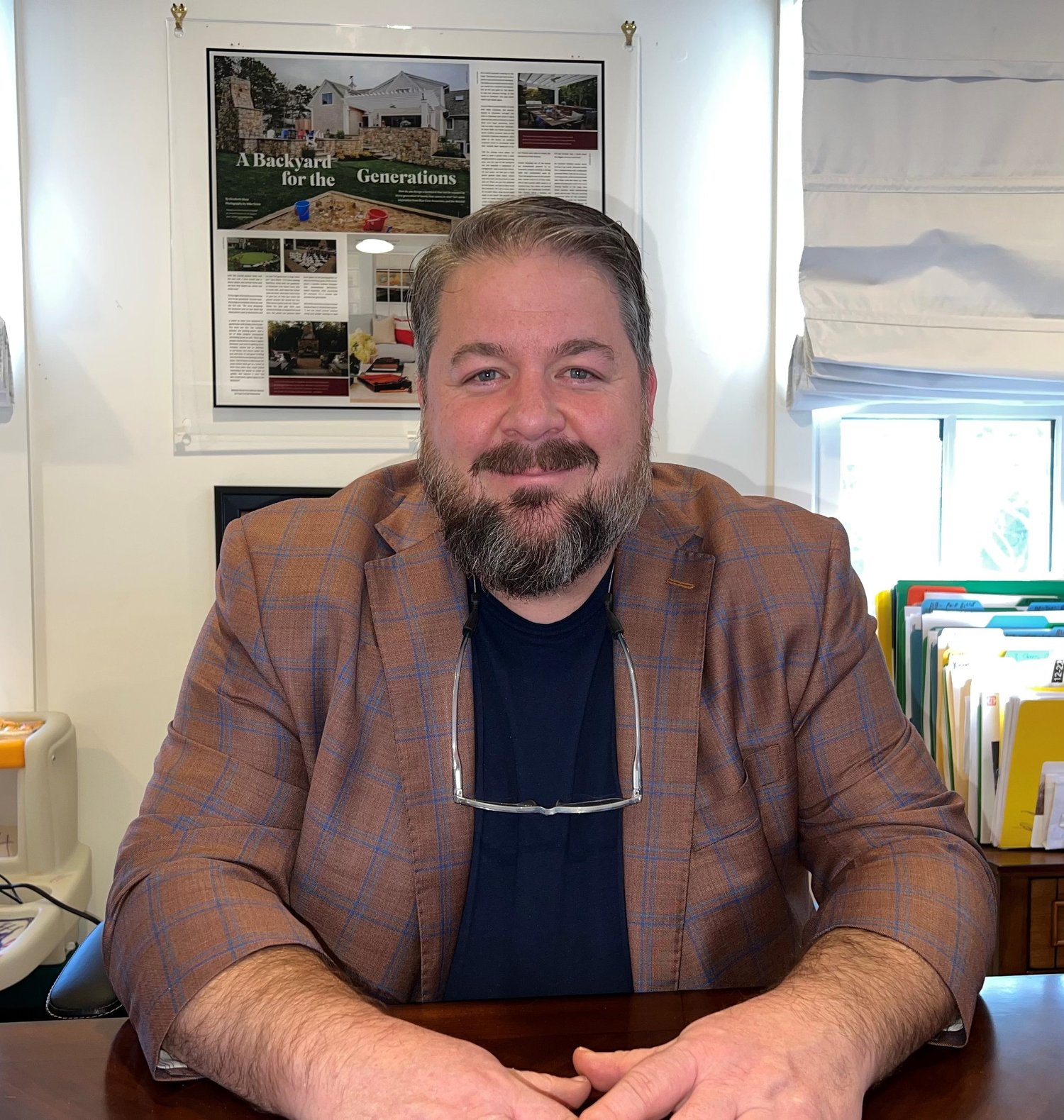 A man with a beard and short hair, wearing a brown plaid blazer and glasses hanging around his neck, sitting at a desk in a well-lit office with a smiling expression.