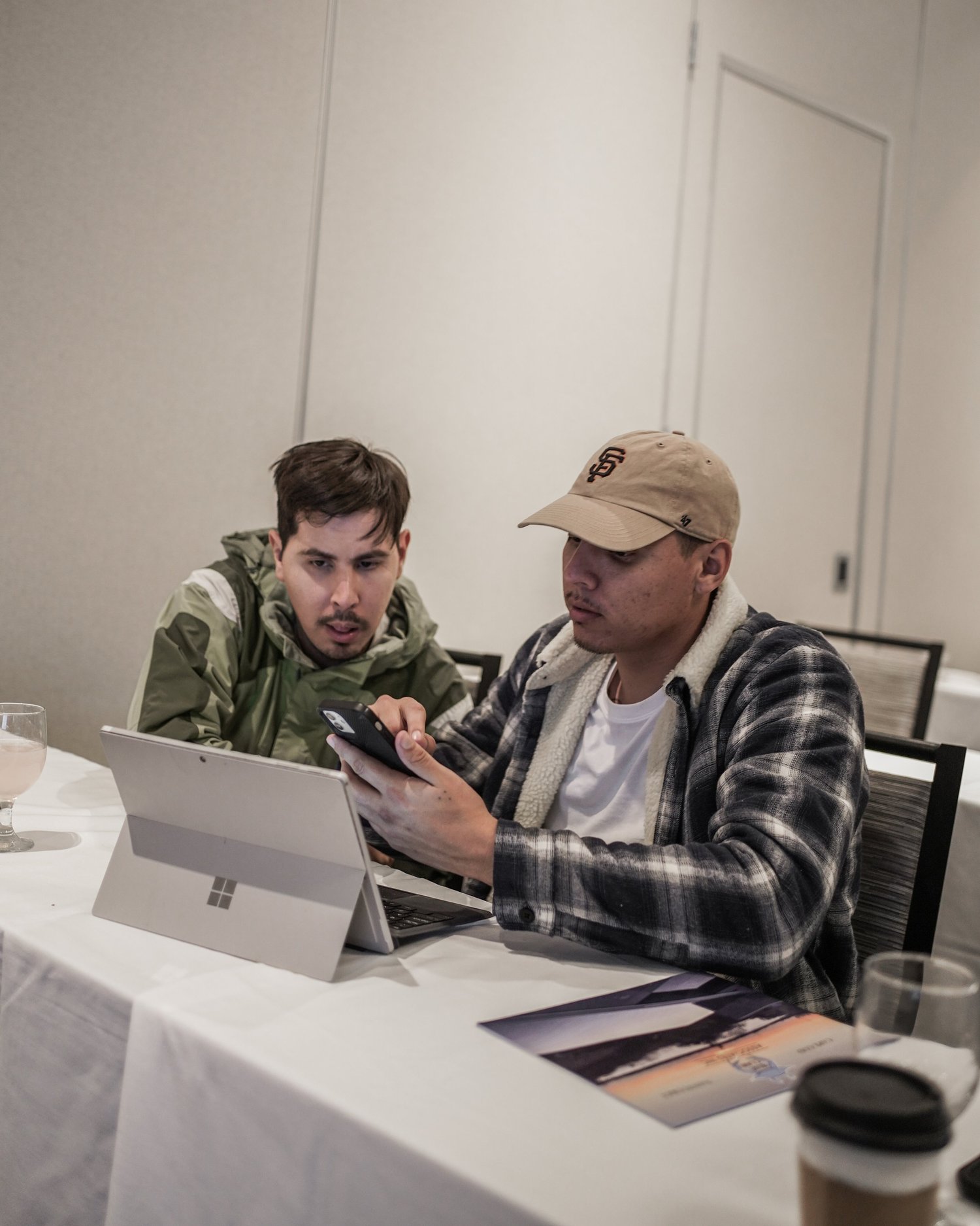 Two young men sitting at a table looking at a phone together, with a tablet, a brochure, a glass of water, and a coffee cup on the table.