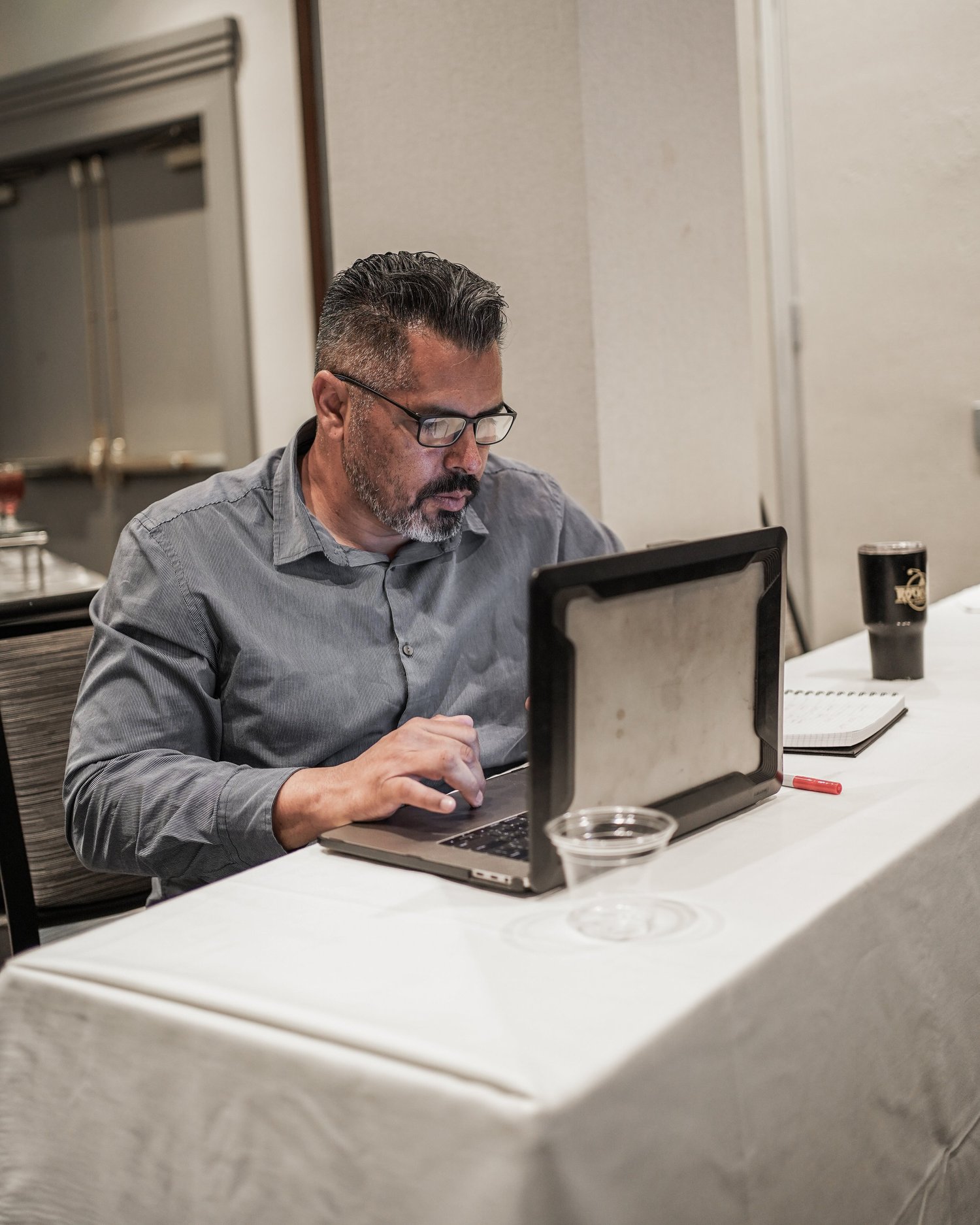 A man with glasses, a gray shirt, and a beard is working on a laptop at a table with a white tablecloth. There is a glass dish and a black cup on the table.