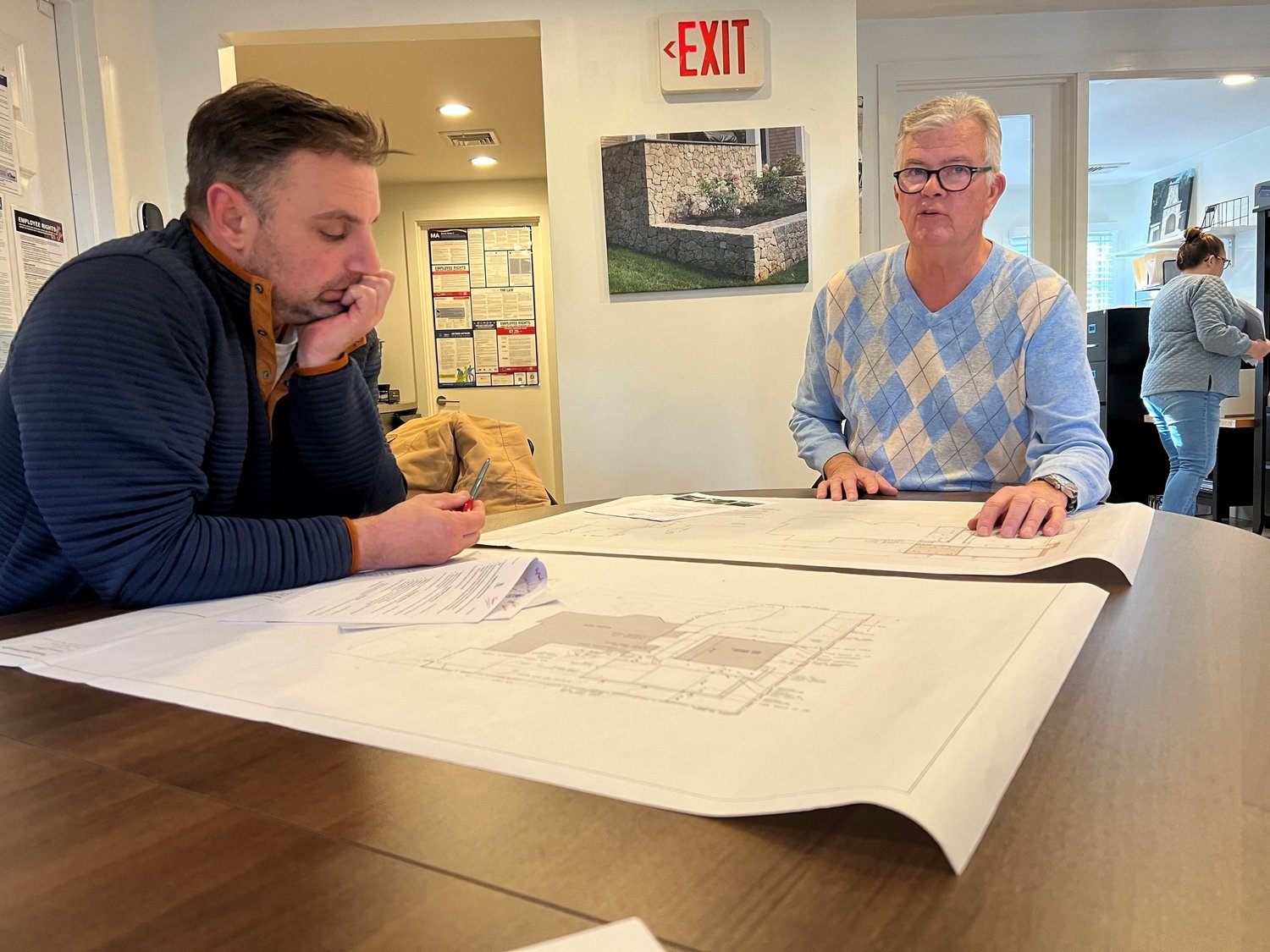 Two men are seated at a table with architectural plans, discussing them. One man is on the left, resting his chin on his hand, and the other man is on the right, standing and pointing to the plans. In the background, a woman is working at a counter, 