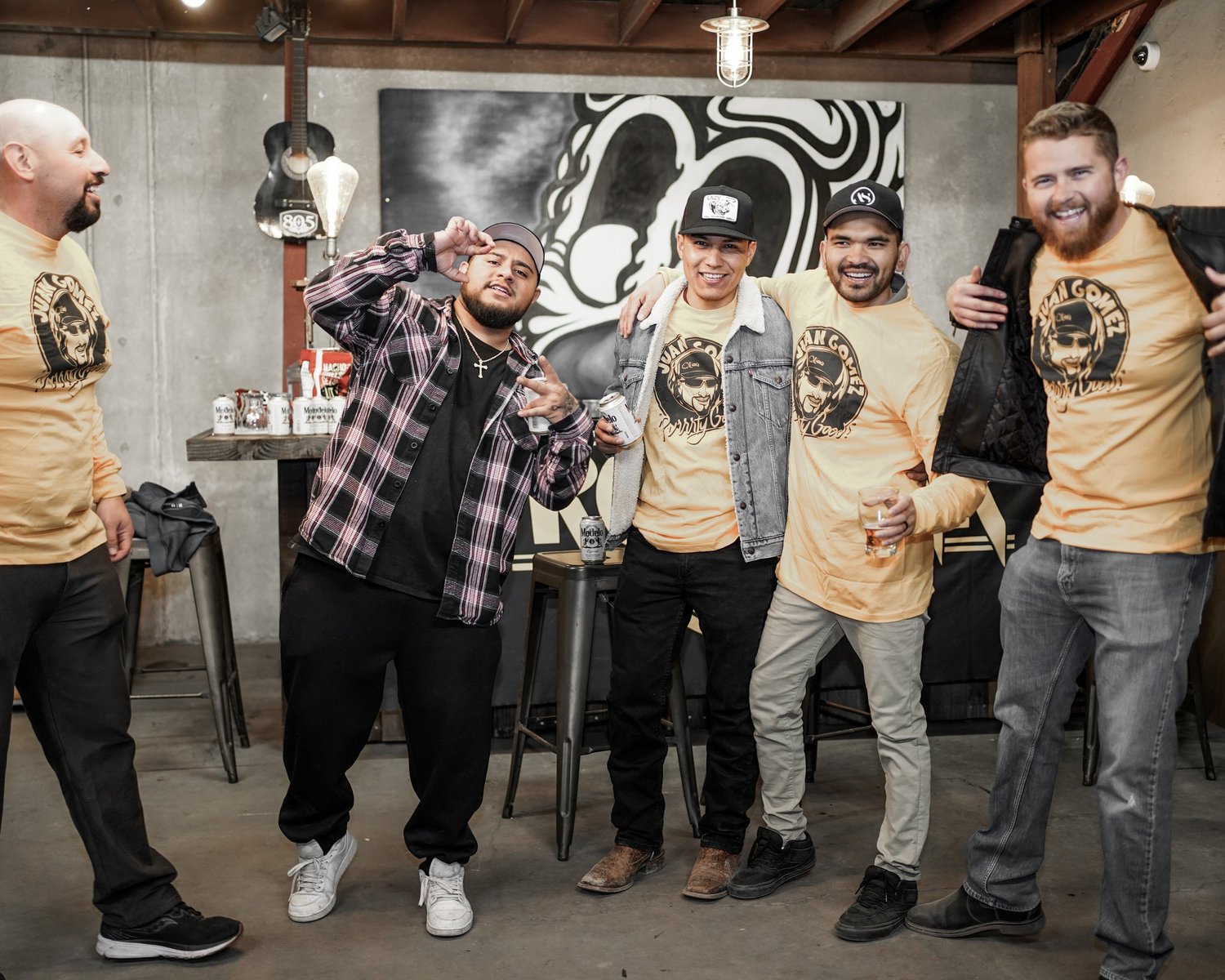 Group of six men smiling and posing together in a bar or brewery, some holding drinks, wearing casual clothing including yellow t-shirts with a graphic design.