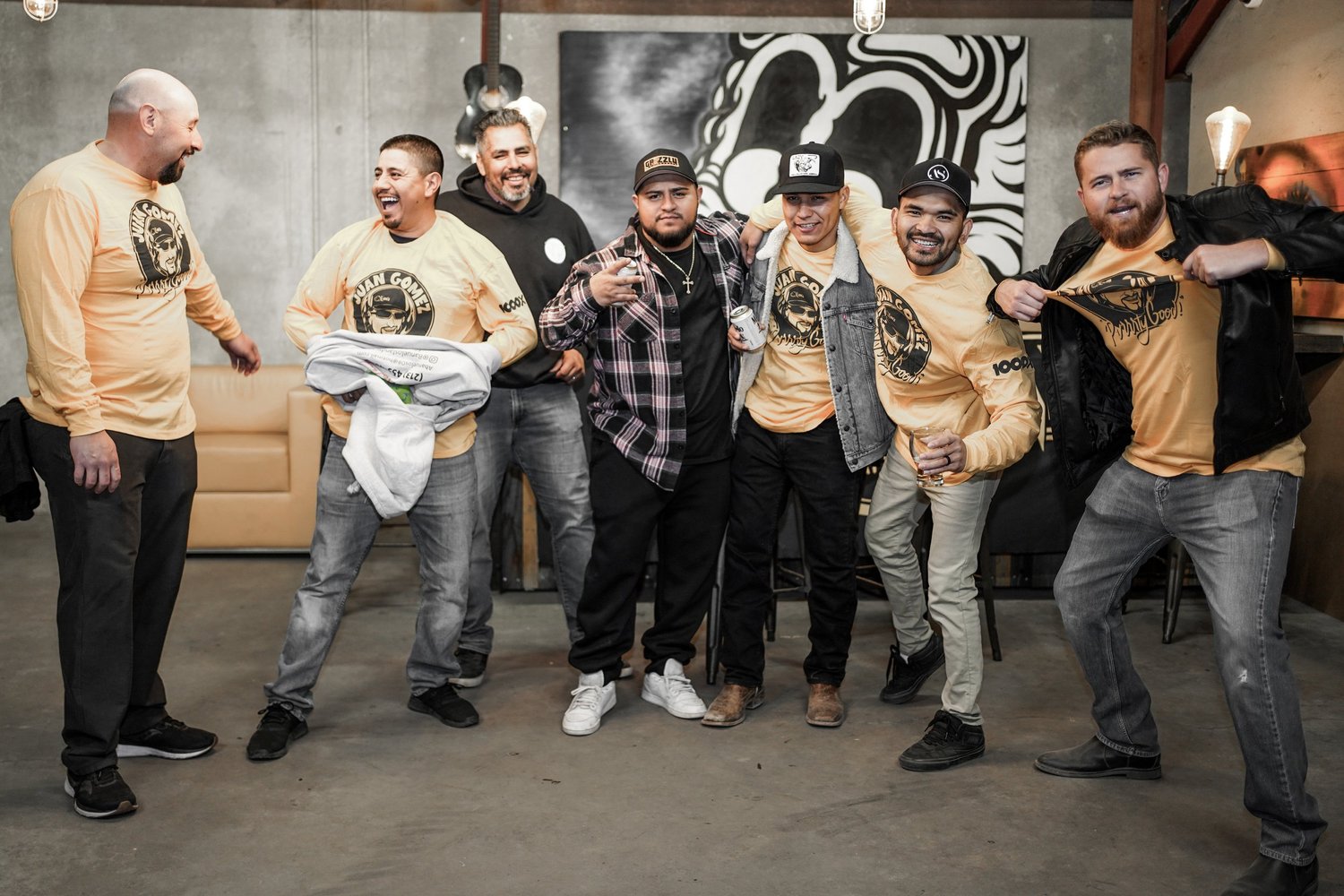 A group of seven men posing together in an indoor setting, some wearing yellow long-sleeve shirts with a logo, one person in black and red plaid shirt, and others in casual attire, smiling and enjoying each other's company.