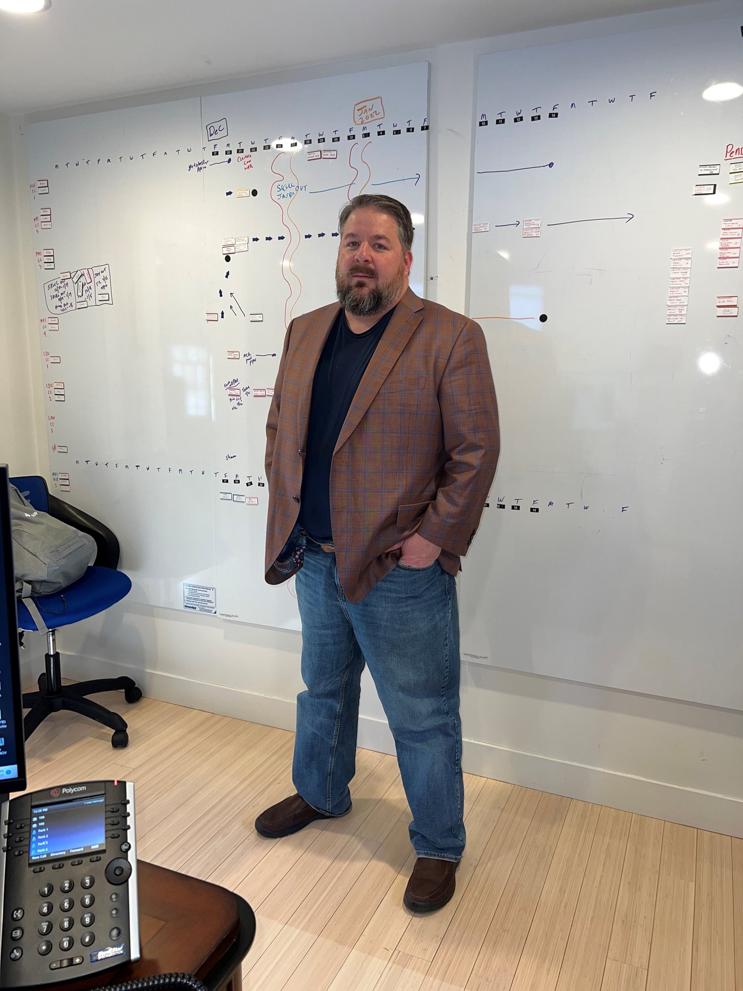 A man with a beard and short hair, wearing a plaid brown blazer, black shirt, jeans, and brown shoes, standing in front of a large whiteboard with various notes and diagrams.