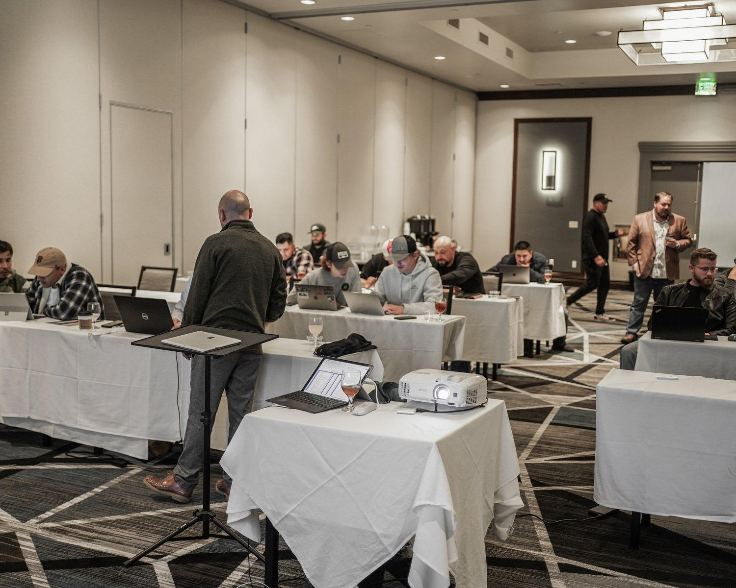 People attending a conference or workshop in a hotel banquet room, sitting at tables with laptops and drinks, while a man stands at a lectern with a laptop and projector.