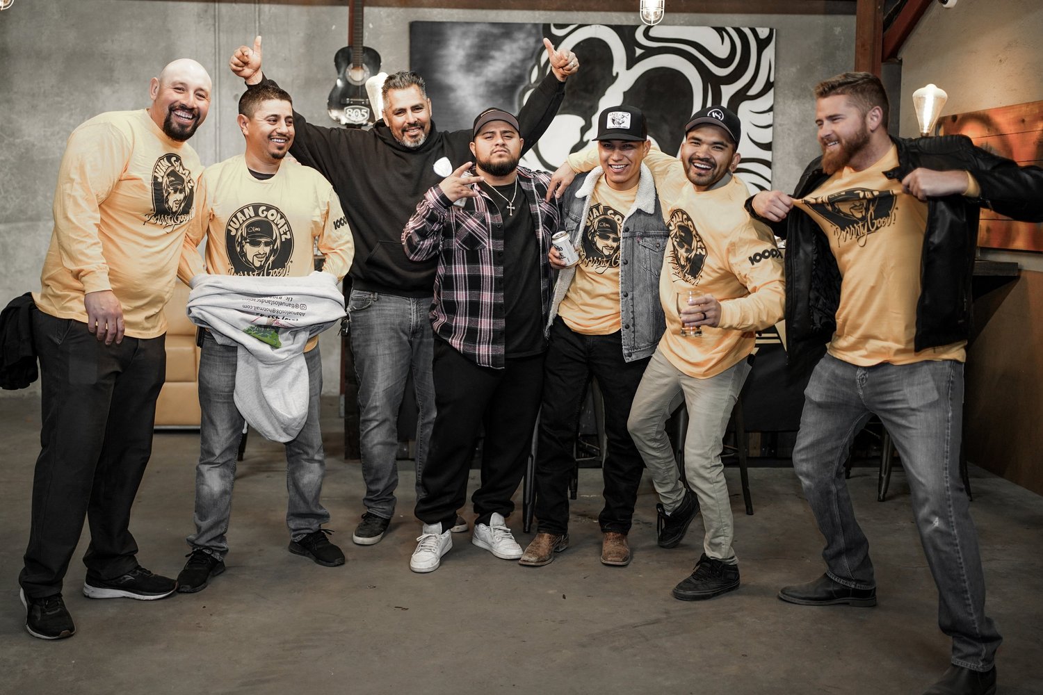 Group of seven men celebrating indoors, some wearing matching yellow t-shirts with a graphic logo, with one man in black hoodie and others in casual jackets, posing cheerfully with drinks and making gestures.
