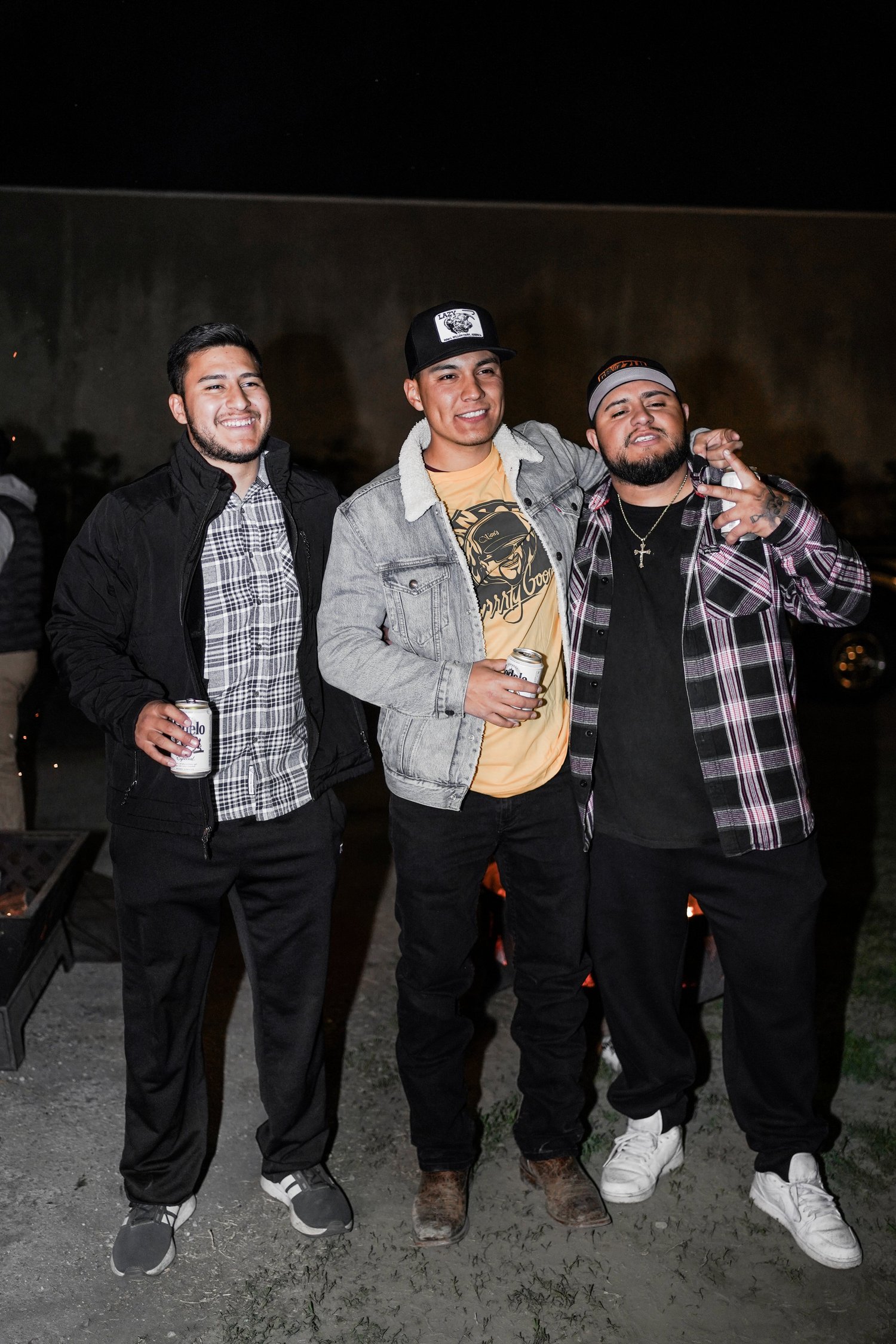 Three young men standing together outdoors at night, smiling and holding drinks.