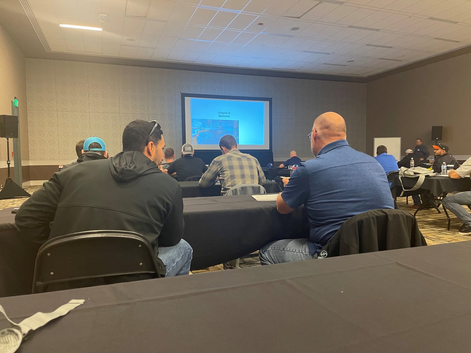 Attendees sitting at tables in a conference room watching a presentation on a large screen.