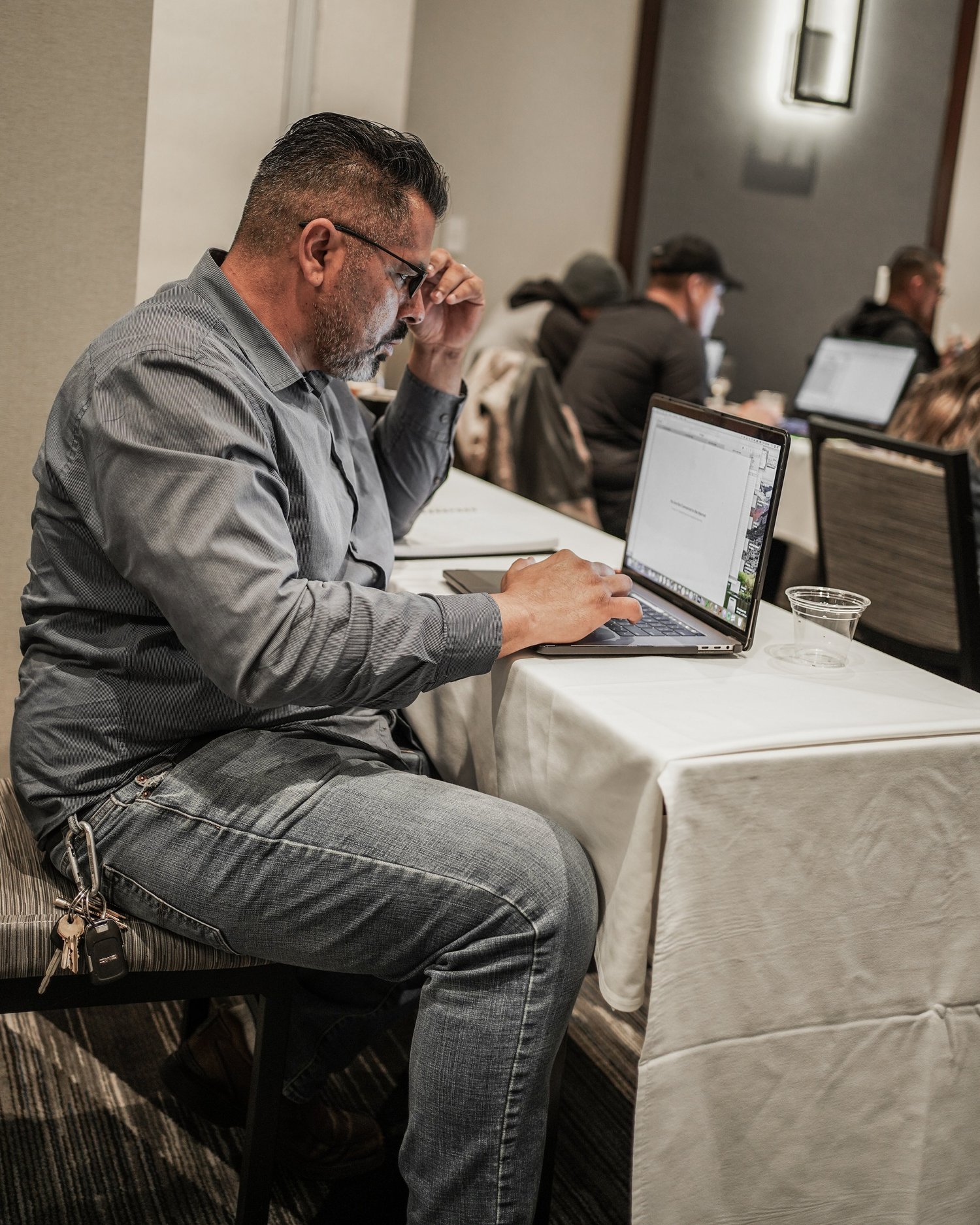 A man sitting at a conference table working on a laptop with other people in the background.