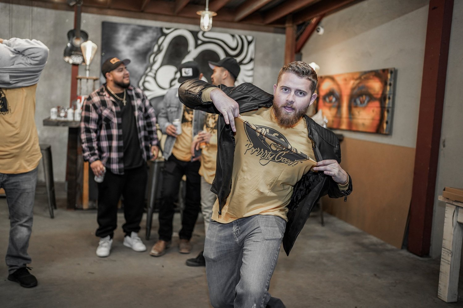 A group of casually dressed men socializing at a bar or brewery in an industrial-style setting with artwork on the walls. One man in the foreground is pulling up his T-shirt to reveal a graphic design as he poses for the camera.