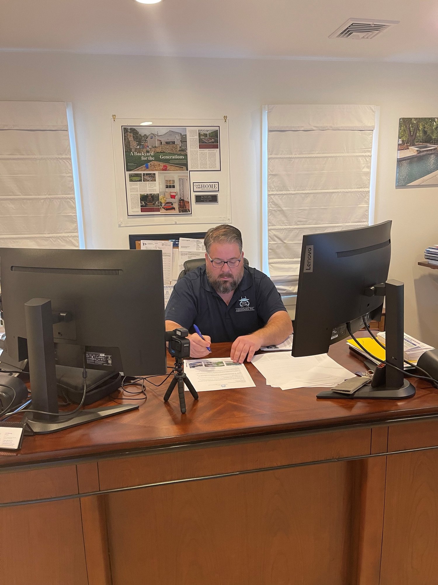 A man with a beard and glasses sits at a wooden desk, surrounded by two large computer monitors, writing on papers with a pen. There is a small tripod with a camera on the desk, and several stacks of papers and files. Behind him are two windows with 