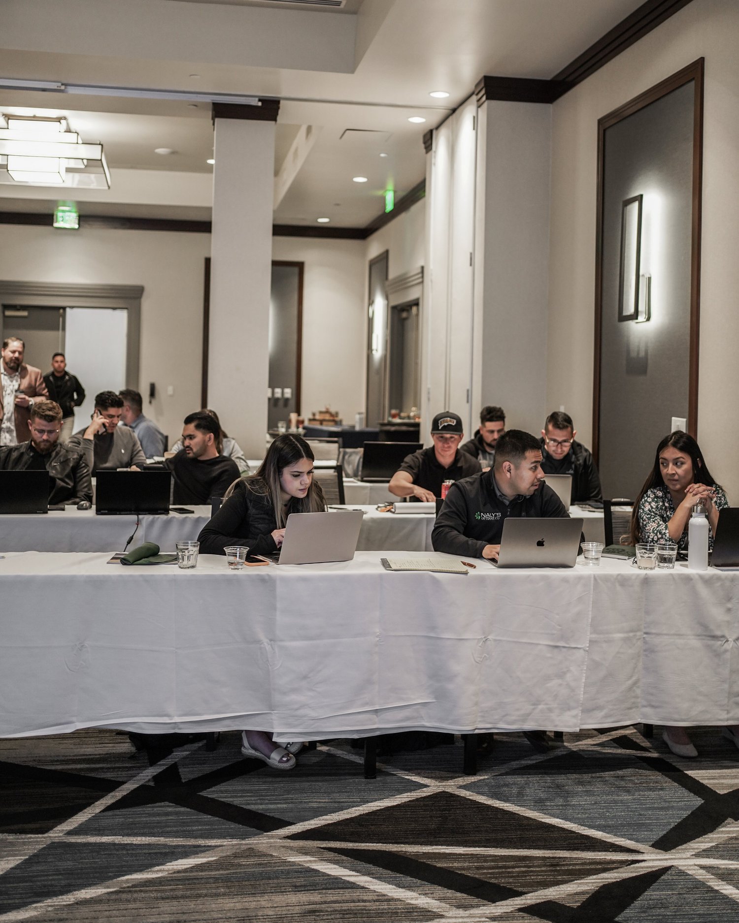 People attending a conference or meeting seated at tables with laptops and notebooks in a large, well-lit room.