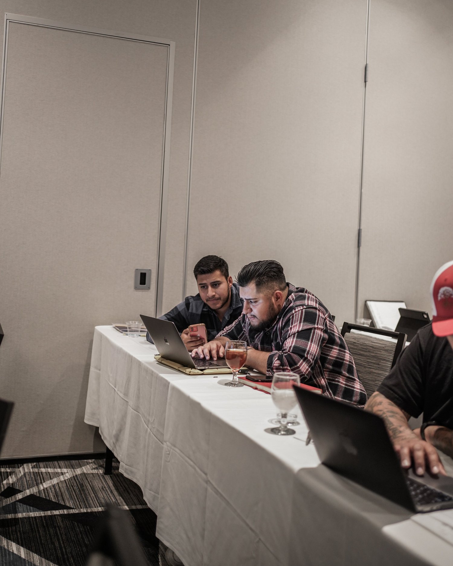 Three men sitting at a conference table, two using laptops, one with a smartphone, with drinks in front of them, in a meeting room.