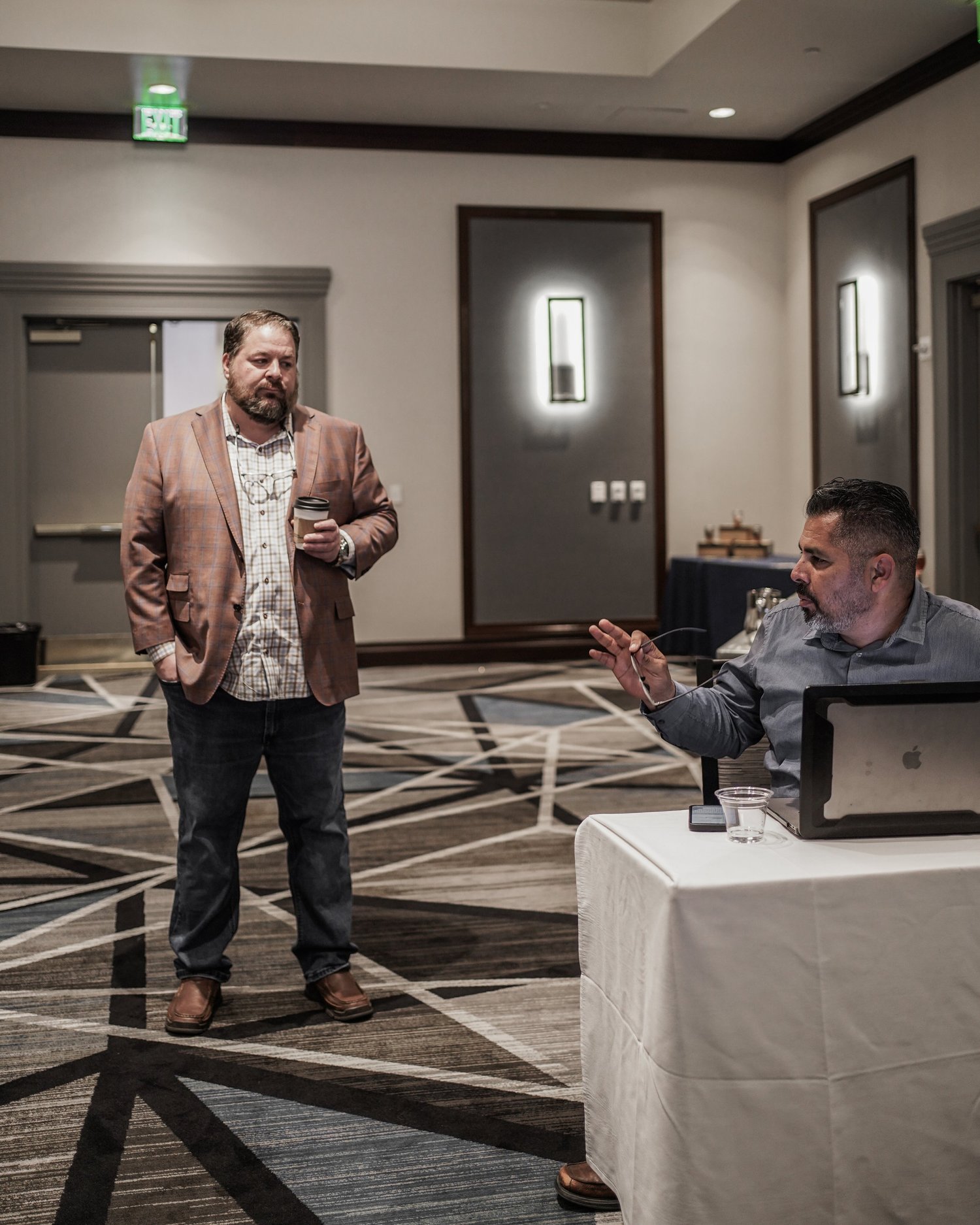 Two men having a conversation in a conference room, one standing with a coffee cup and the other sitting with a laptop and a glass of water.