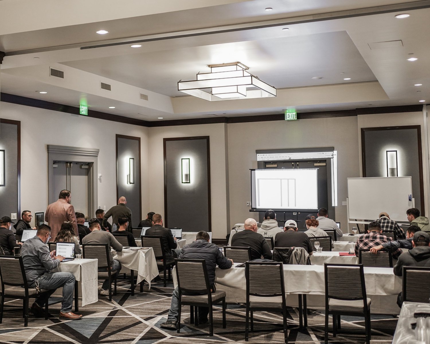 People attending a conference or seminar in a hotel banquet room with presentation screens, whiteboards, and seating arrangements.