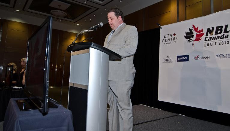 A man in a light gray suit stands at a podium, speaking into a microphone at a formal event, with a large banner behind him displaying logos for GTA Center, NBL Canada, and sponsors.