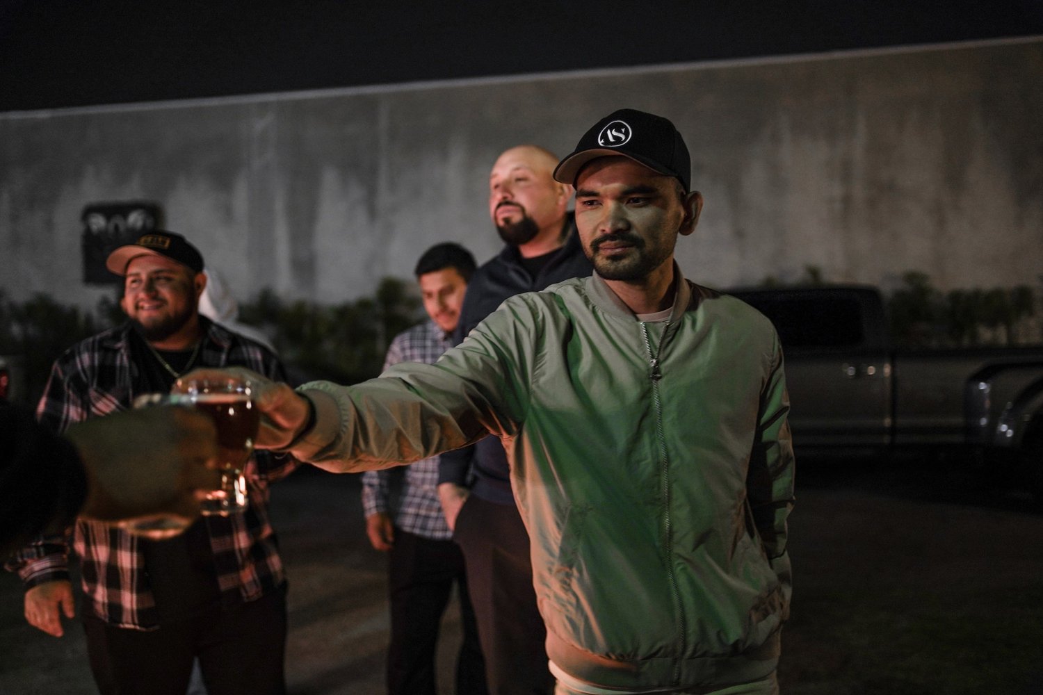 A group of men standing outdoors at night, one man in the foreground extending a glass of beer to another person, others smiling and watching.