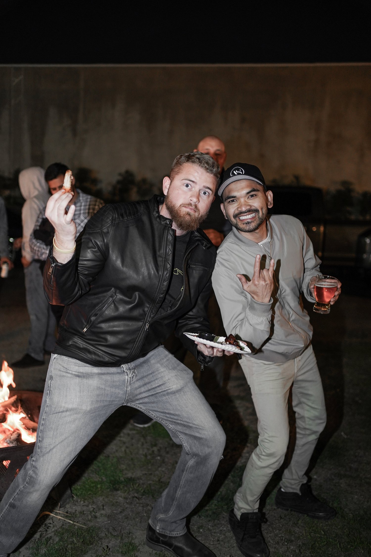 Two men at an outdoor night gathering, one holding a plate of food and the other holding a glass of drink, posing for the camera, with a fire pit in the foreground.