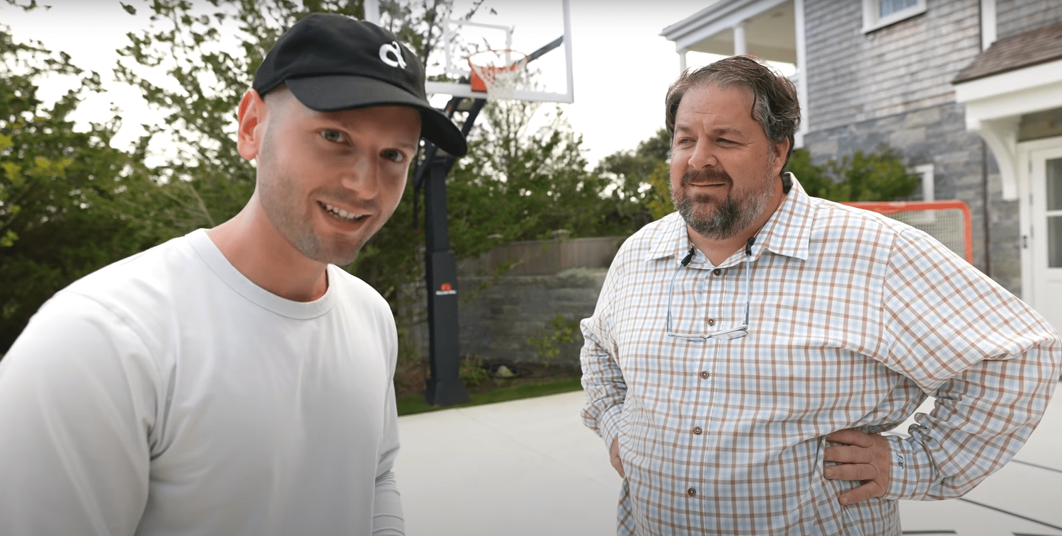 Two men standing outside on a driveway near a basketball hoop, one smiling and the other looking puzzled, with a house and trees in the background.