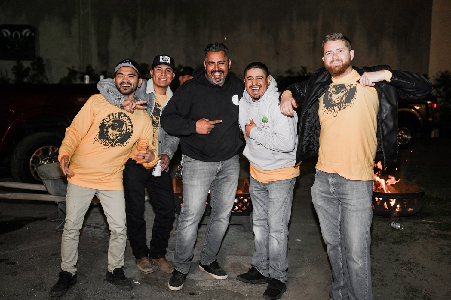 Group of five men smiling and posing for a photo in front of a firepit outdoors at night. Some are wearing matching yellow T-shirts with a logo that says 'Juan Gomez.'