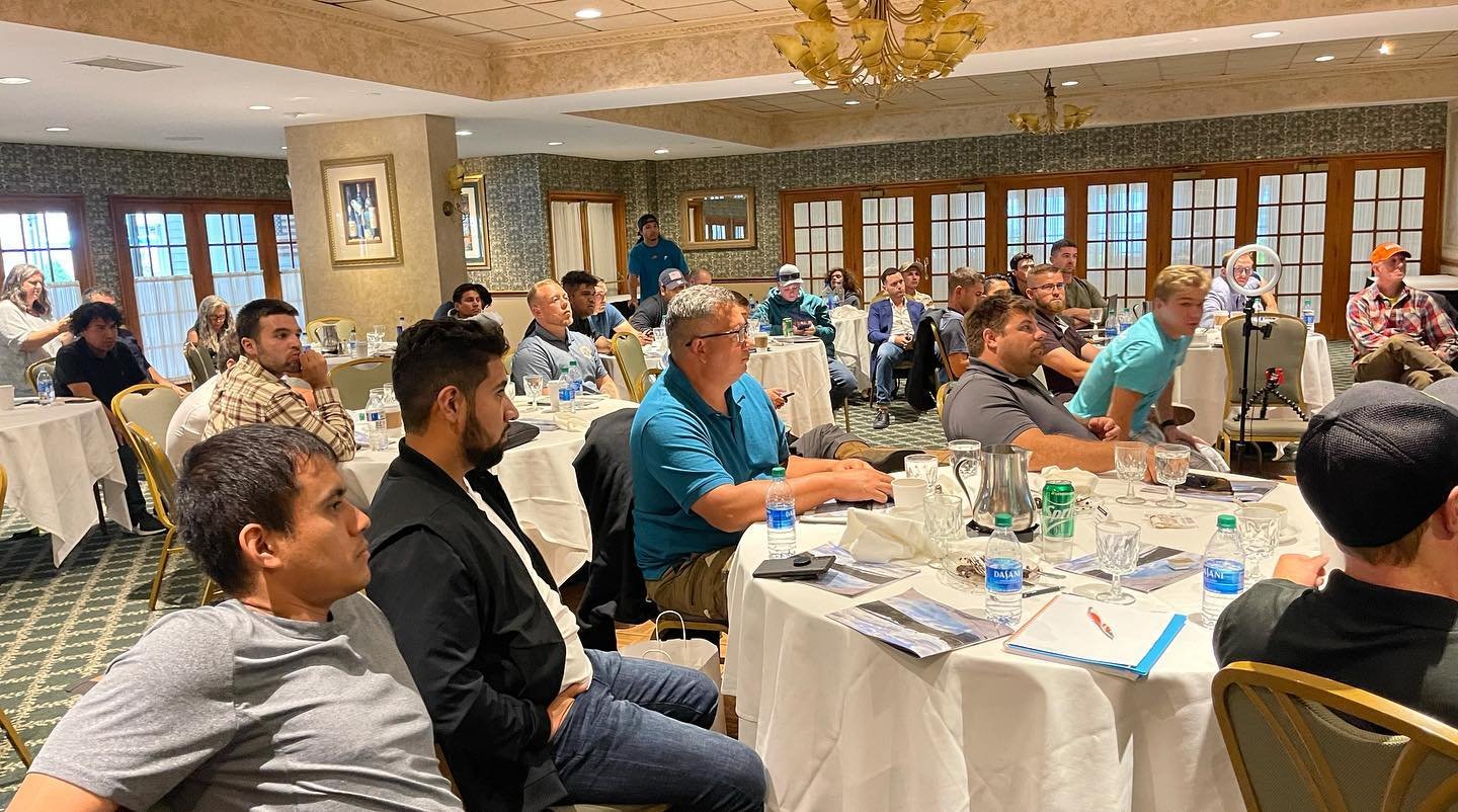 People attending a conference or seminar in a banquet hall with round tables, water bottles, and notebooks.