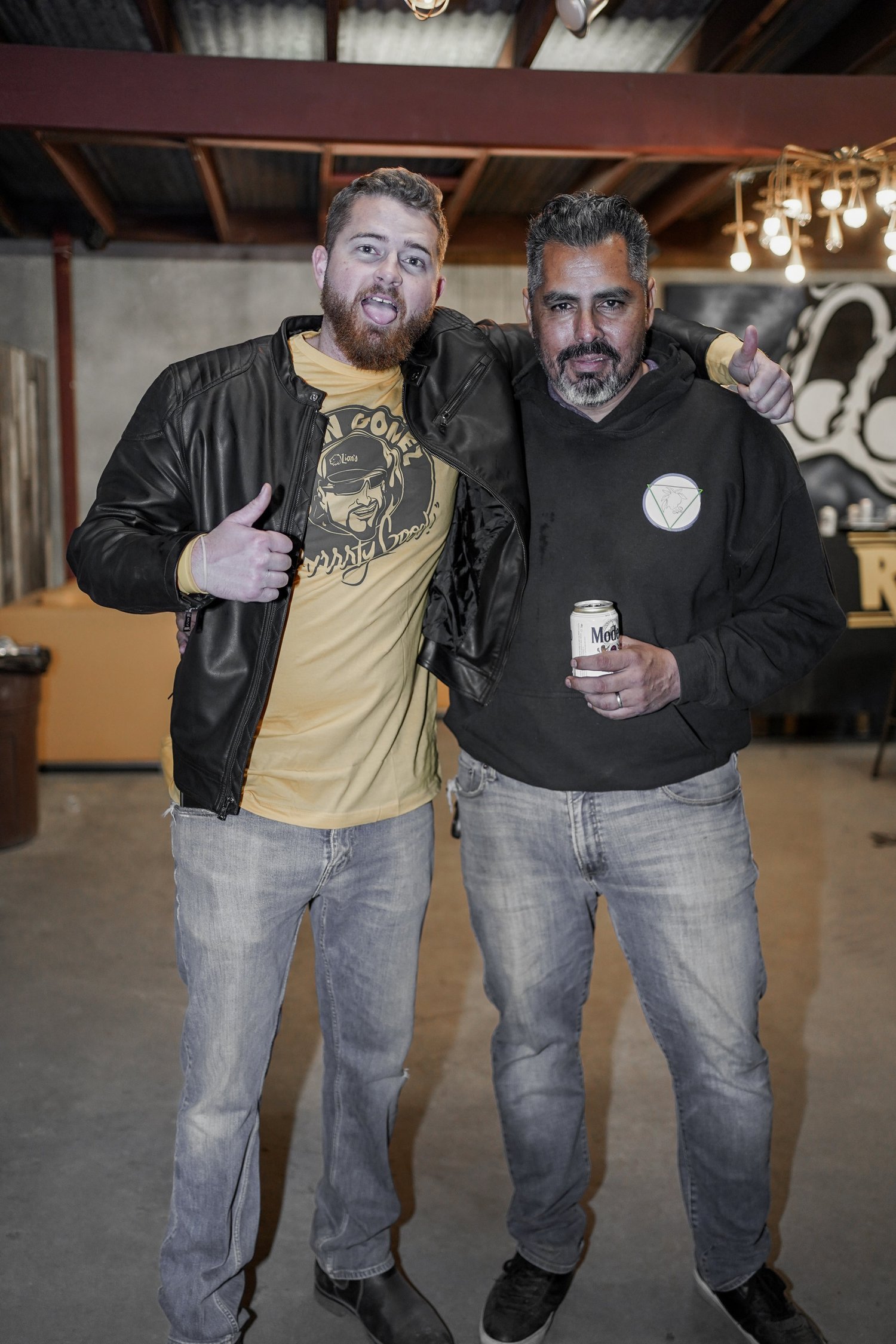 Two men standing together in a garage or industrial space, smiling and posing for the camera. The man on the left has a beard, is wearing a black leather jacket over a yellow graphic t-shirt, and giving a thumbs-up. The man on the right has gray hair
