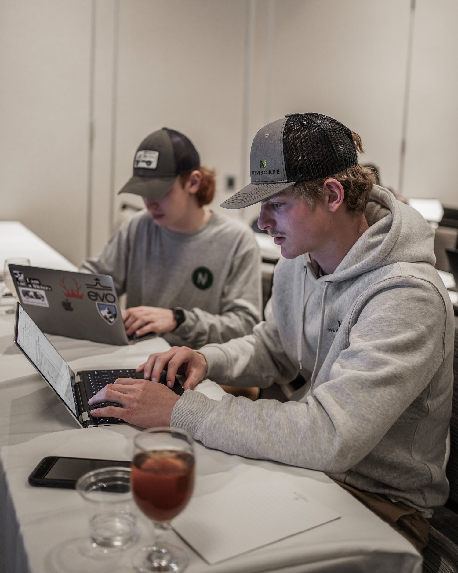 Two young men working on laptops at a table, wearing casual hoodies and baseball caps, with a drink and a phone on the table.