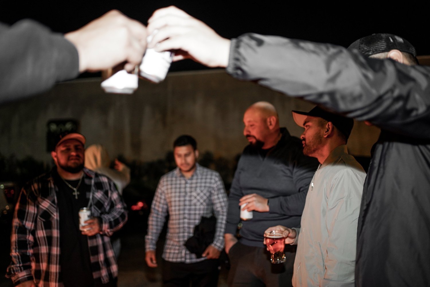 Group of men socializing outdoors at night, holding drinks, with hands passing a shot glass over the scene.