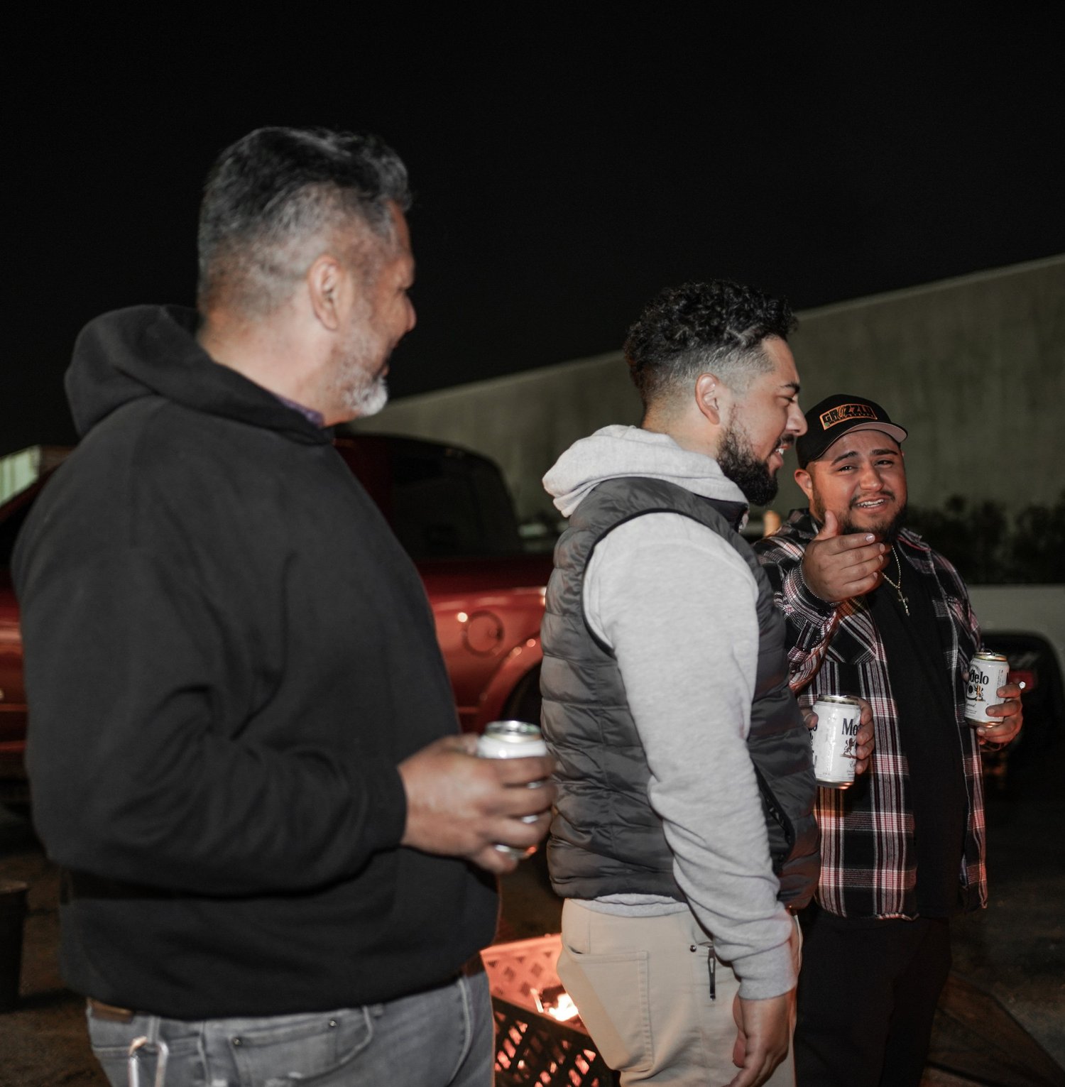 Three men standing outdoors at night, holding cans of Modelo beer, engaged in conversation, with a red truck and a concrete wall in the background.