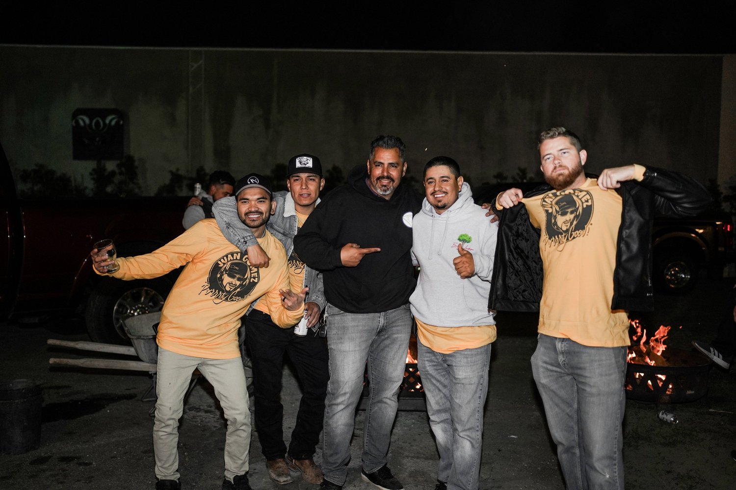 Group of five men smiling and posing together outdoors at night, with a fire pit in the background.