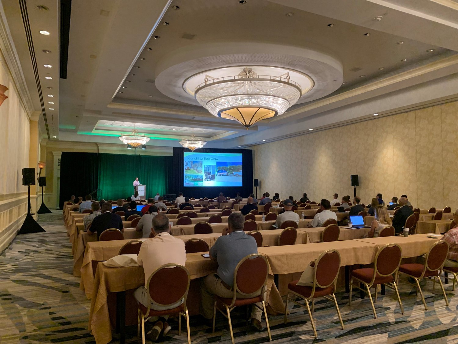 Conference room with attendees sitting at tables, facing a speaker on stage with a projected presentation.