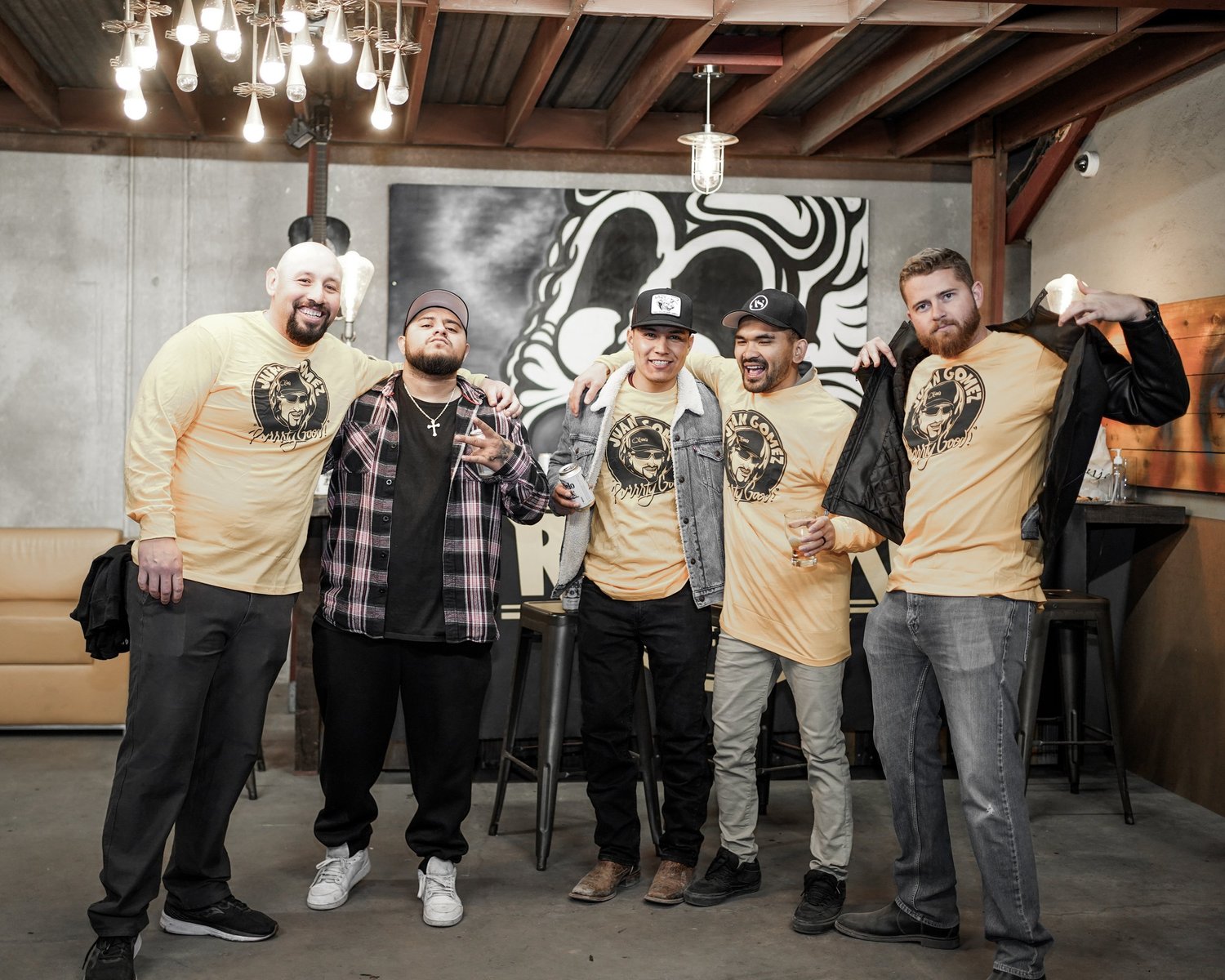 Five men smiling and posing together at a bar or restaurant. They are wearing casual and stylish clothing, with some holding drinks. The background features a black and white mural and modern lighting fixtures.