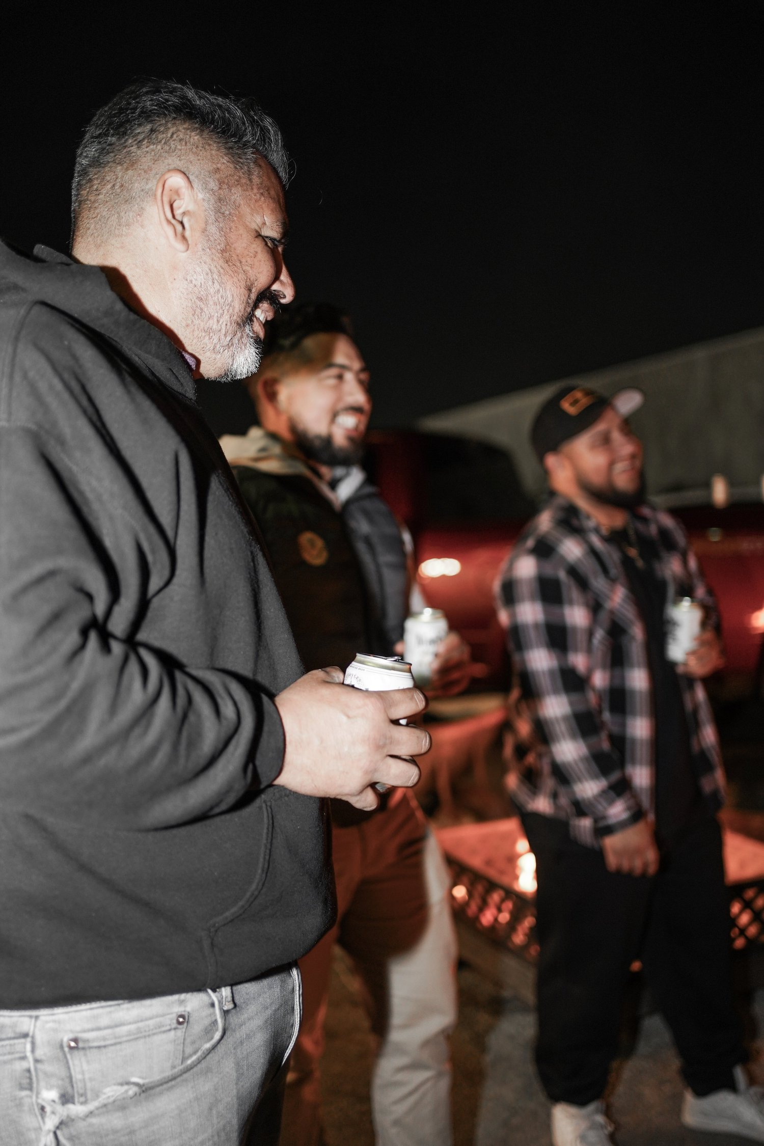Group of men standing outdoors at night, smiling, holding canned drinks, enjoying conversation.