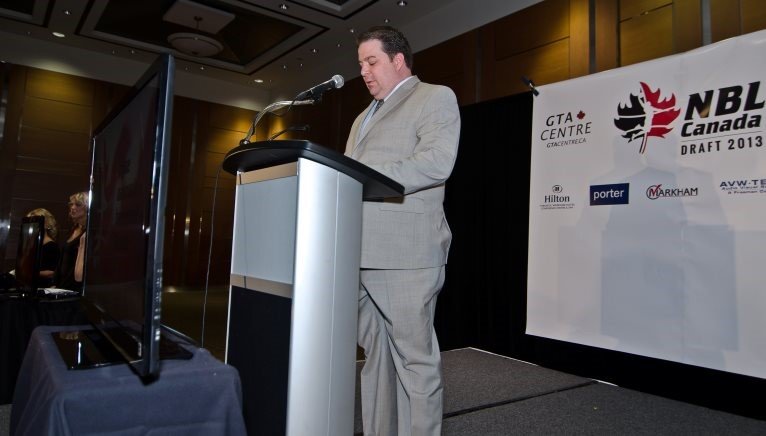 Man in gray suit speaking at a podium during a conference with a banner in the background displaying logos of NBL Canada and other sponsors.