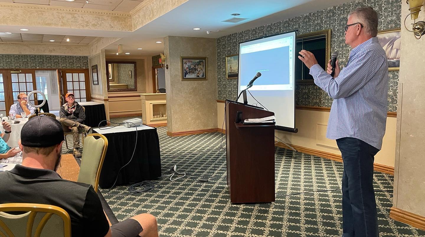 A man giving a presentation in a hotel conference room with a projector screen, presenting to a seated audience.