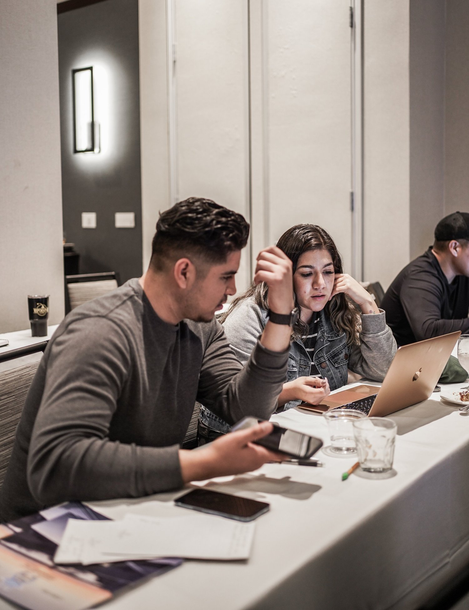 Three young adults sitting at a table in a meeting room, working on laptops and with papers and drinks in front of them.