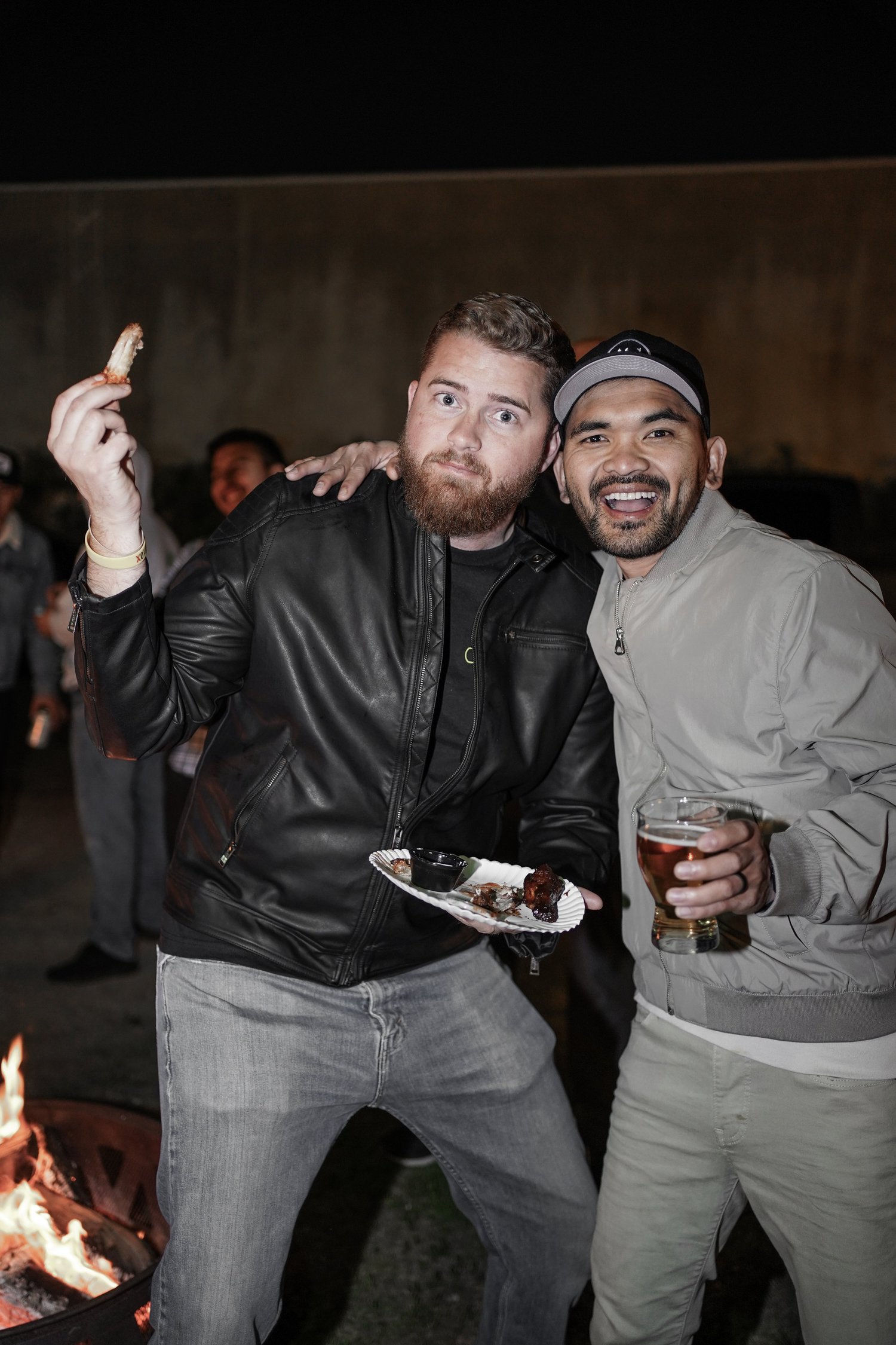 Two men smiling at a nighttime outdoor gathering near a fire pit, one holding a plate with food and a drink, the other holding a drink, with other people in the background.