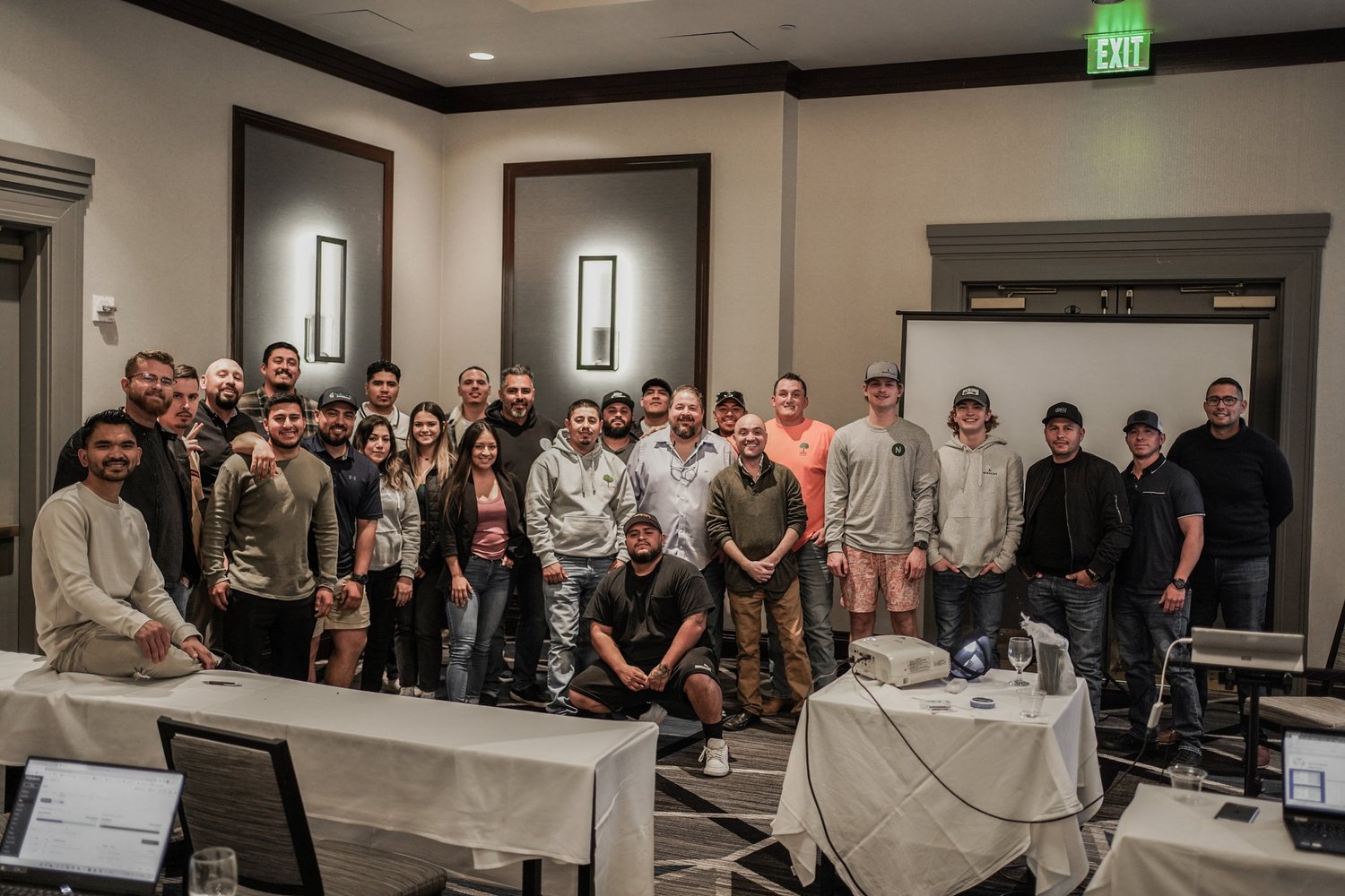 Group of people posing for a photo in a conference room, some standing and some seated, with a white projection screen, tables, and laptops in the foreground.