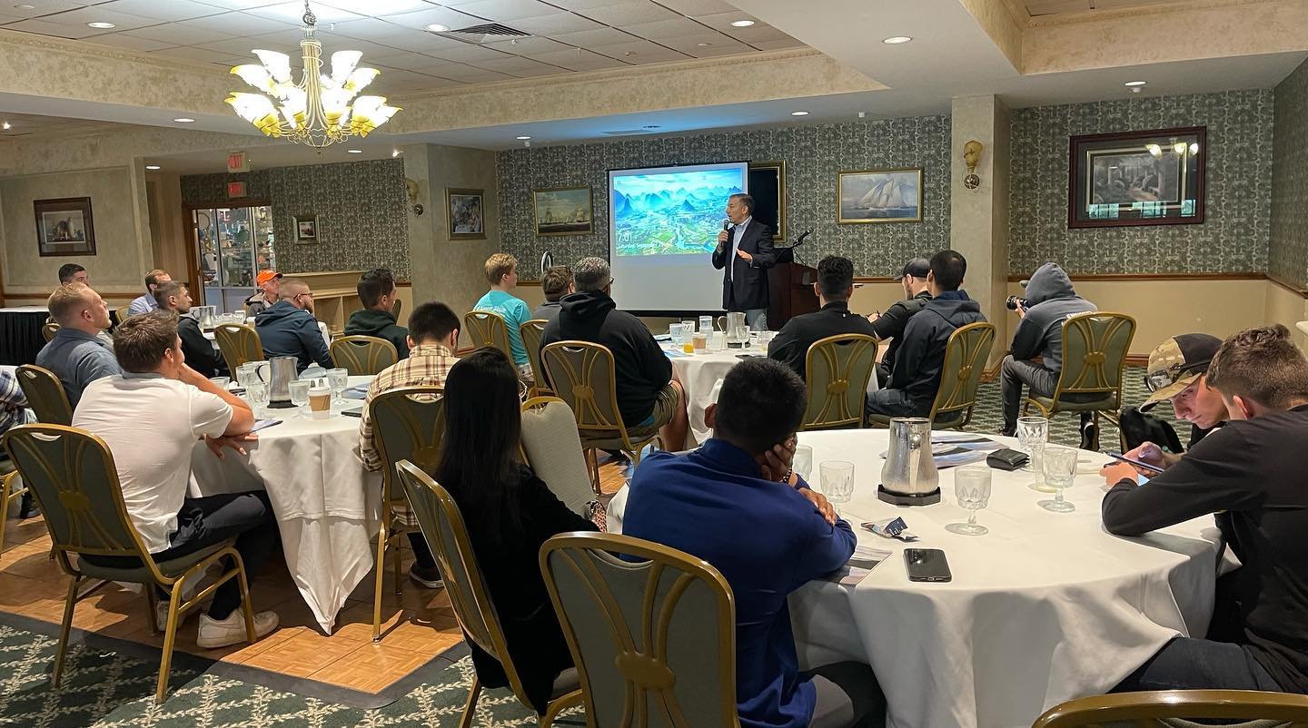 A man in a suit giving a presentation to a group of people seated at round tables in a banquet hall. The presentation screen displays a scenic landscape. Attendees are listening and some are taking notes or using phones.